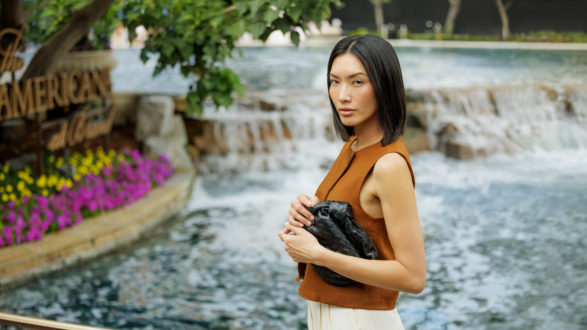 Model in rust top holding woven clutch by waterfall at The Americana at Brand
