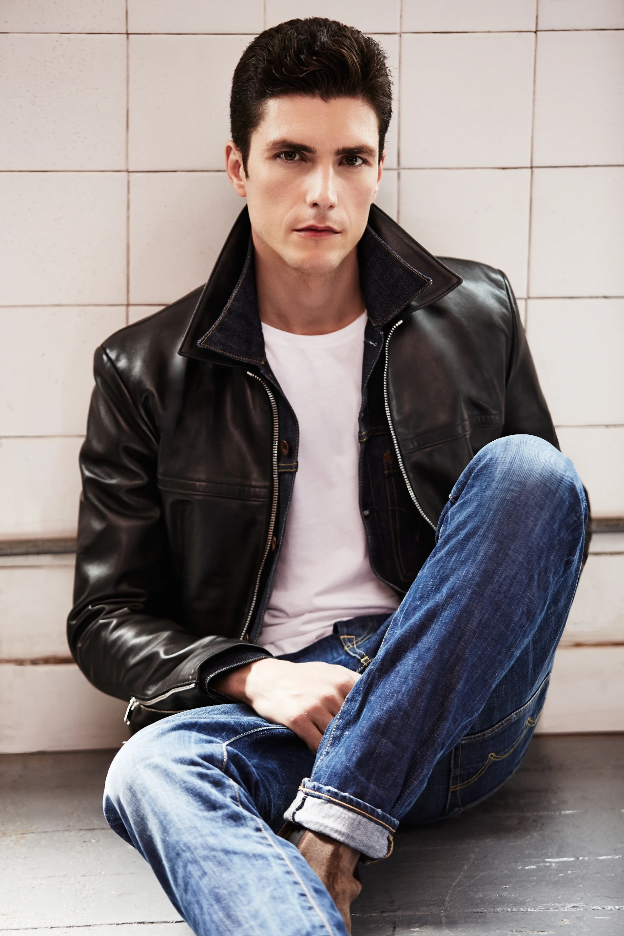 Man in black leather jacket and blue jeans sitting against white tiles