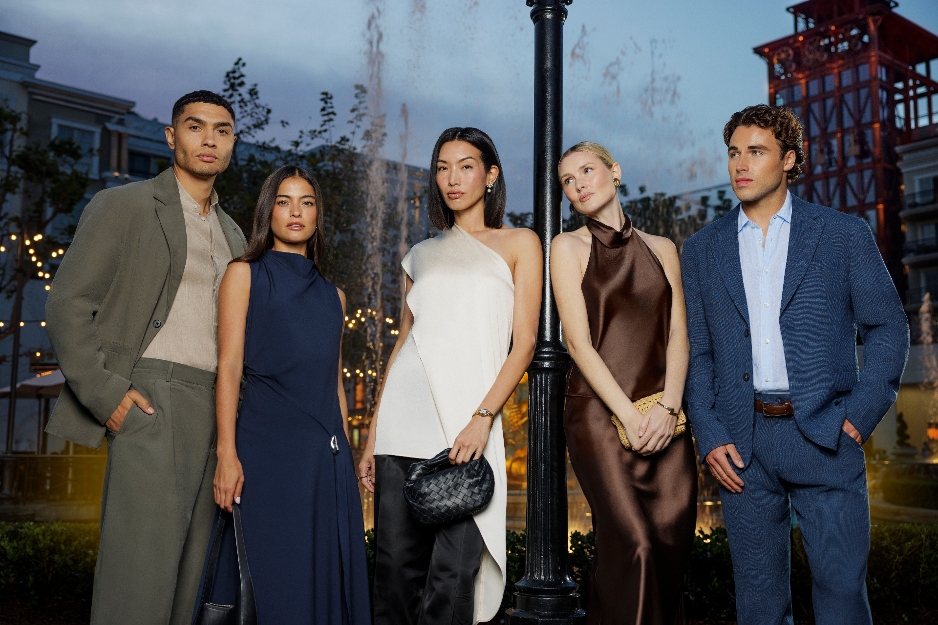 Five models in evening wear standing outdoors at dusk beside a black lamppost